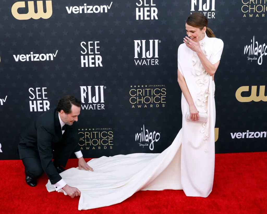 Matthew Rhys, in a black tuxedo, kneels to adjust the train of Keri Russell's white gown on the red carpet.