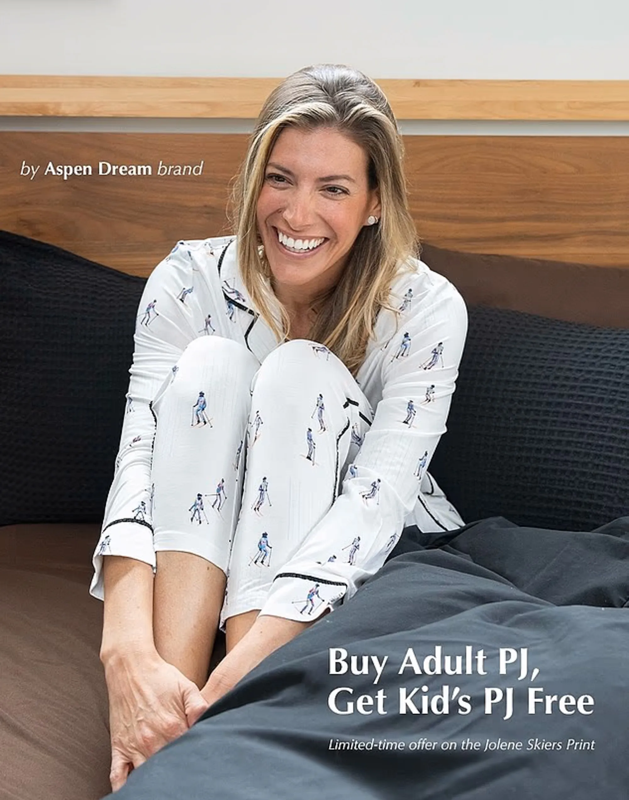 A smiling woman wearing white pajamas with a skier print sits on a bed with dark bedding.
