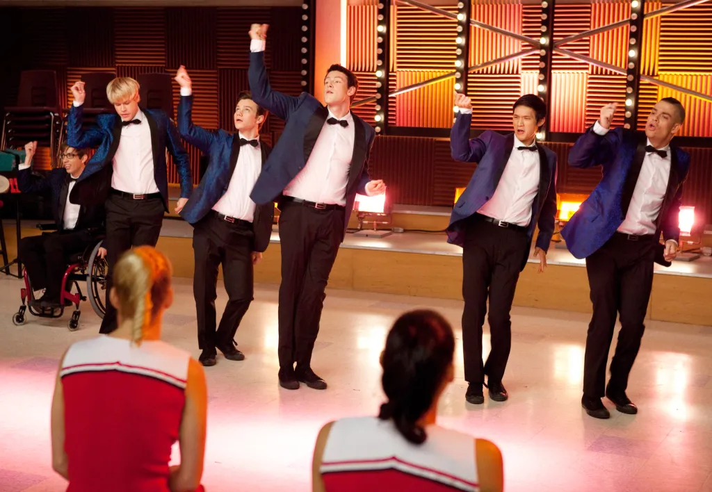 Six male singers from the TV show Glee in blue and black tuxedos, performing and snapping their fingers, with two cheerleaders watching them in the foreground.