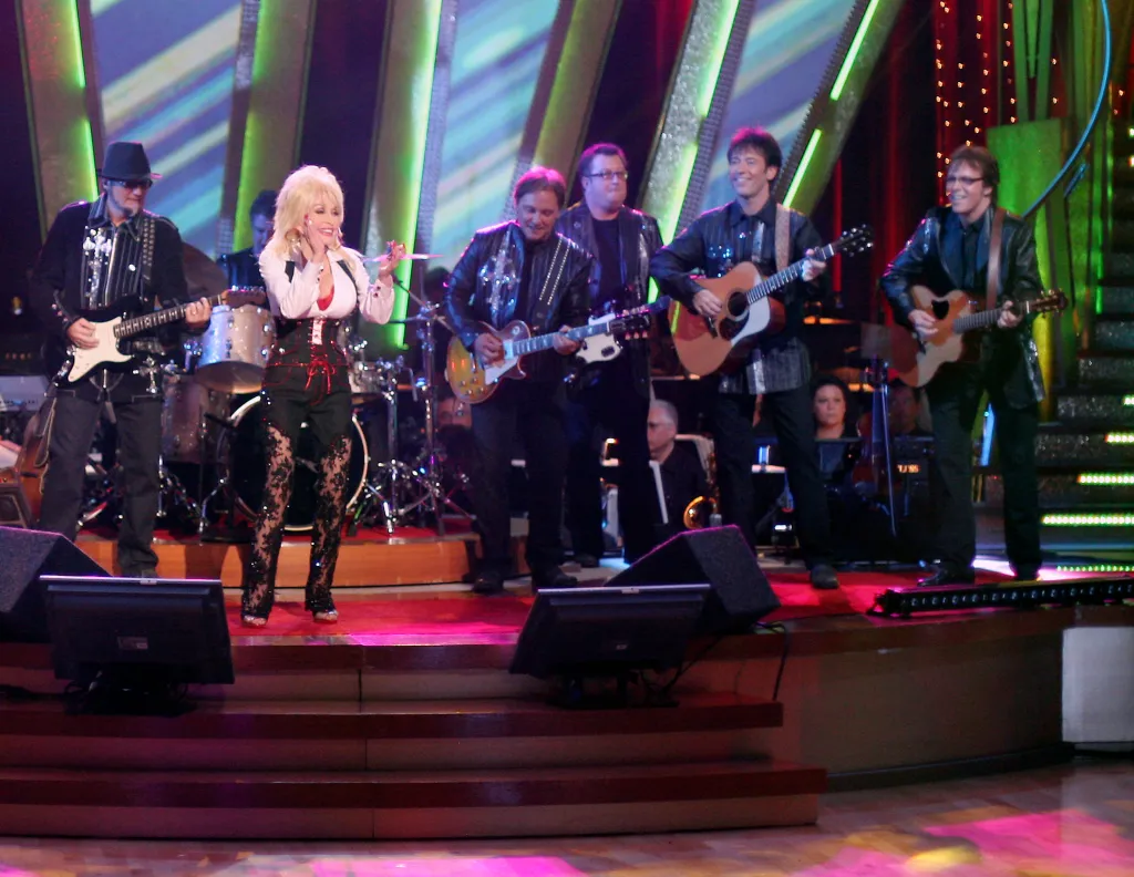 Dolly Parton singing with her band, dressed in a white shirt and black lace pants.