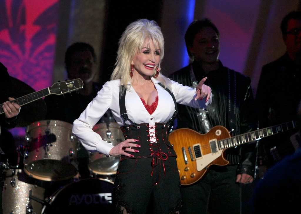 Dolly Parton performing on stage in a white shirt, red corset, and black pants, pointing towards the audience.