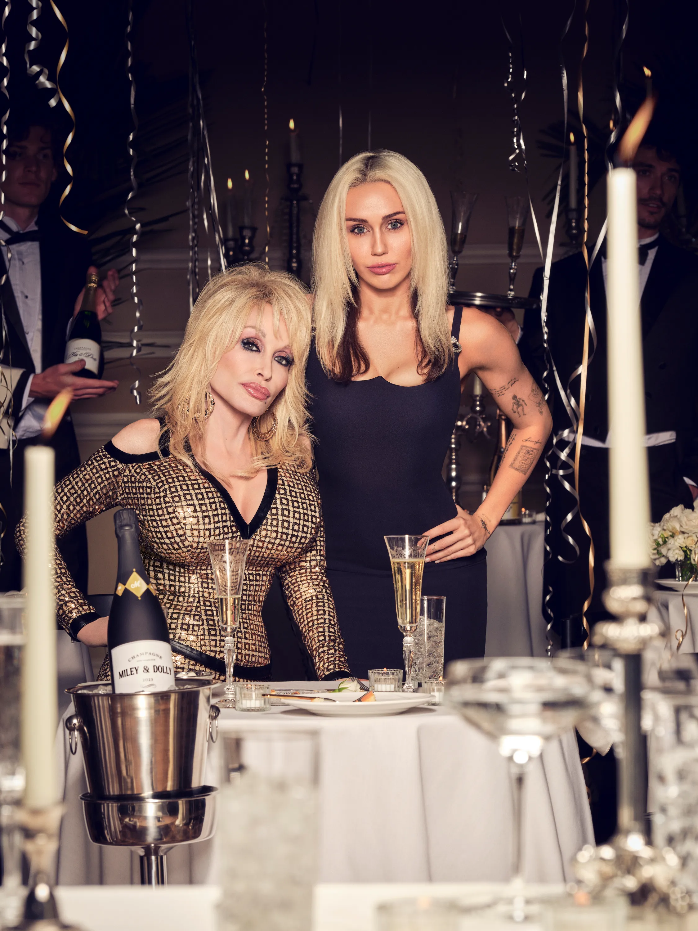 Dolly Parton in a gold sequined dress and Miley Cyrus in a black dress stand behind a table with champagne and candles.
