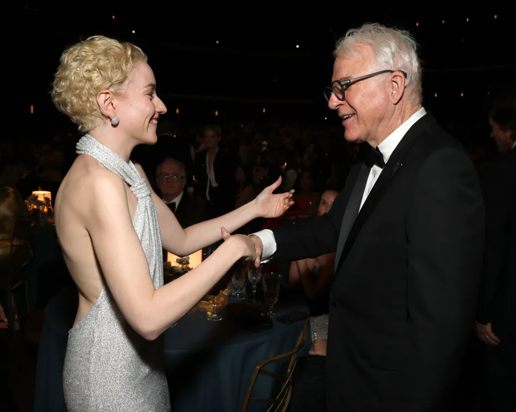 Julia Garner, in a shimmering silver gown, smiles at Steve Martin, who extends his hand towards her at the 78th Primetime Emmy Awards.