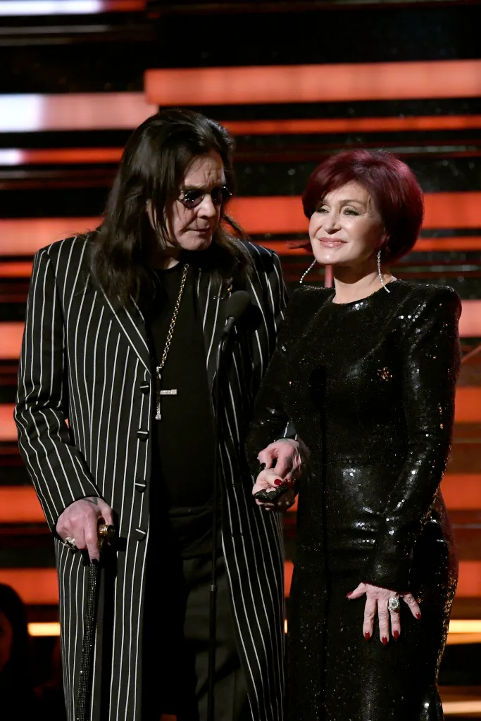 Ozzy Osbourne wearing a striped suit and sunglasses, holding Sharon Osbourne's hand while she wears a black sequined dress.