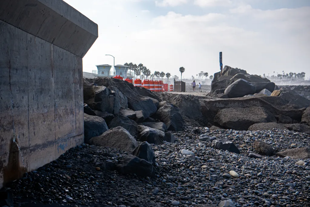 Tamarack State Beach in Carlsbad, California, has been damaged by large waves, leaving a rocky, eroded landscape with a concrete barrier, caution barrels, and distant lifeguard towers.