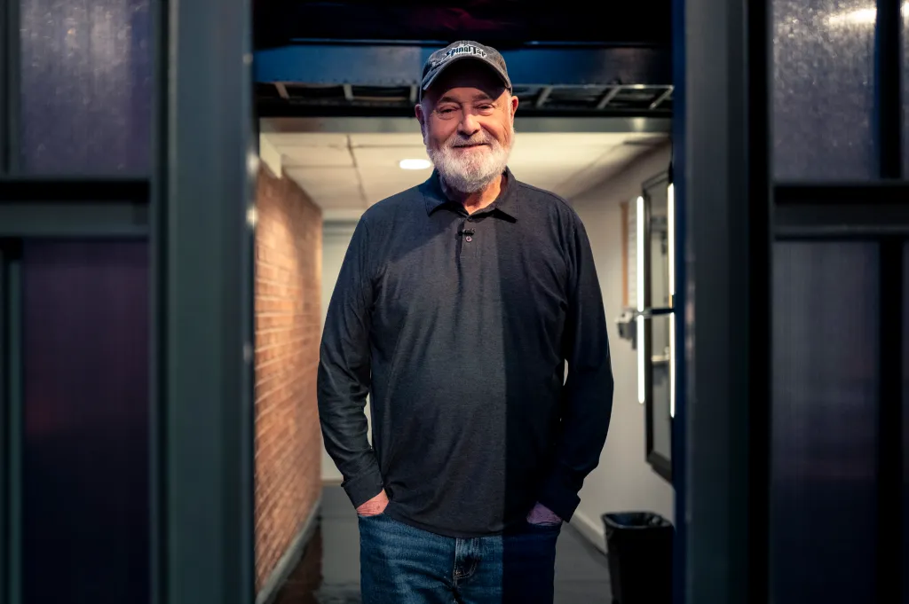 Director Rob Reiner smiles while wearing a baseball cap and dark shirt.