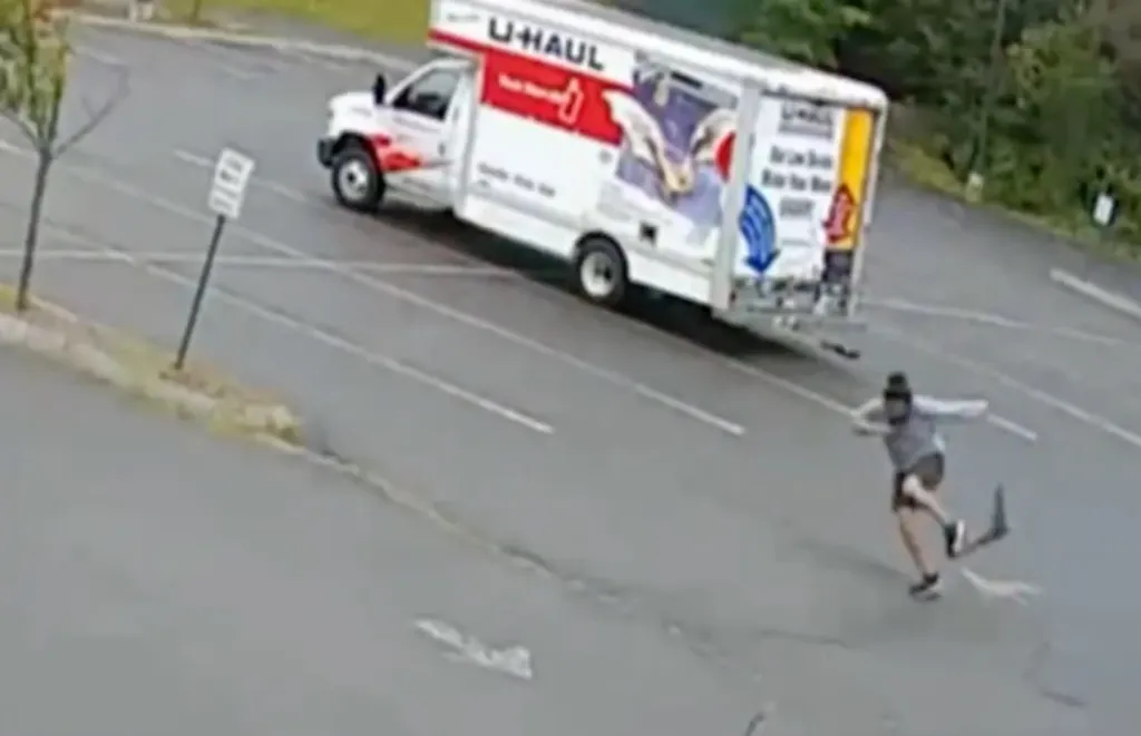 A U-Haul truck drives past a person running away from a fisher, which is a weasel-like animal.
