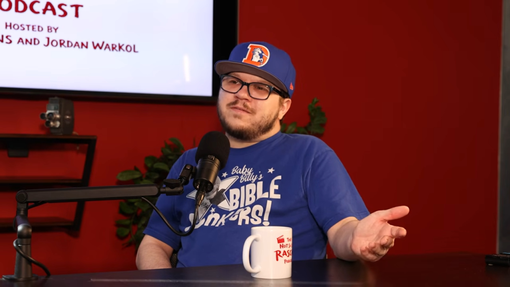 Justin Cooper, former child star of Liar Liar, wearing a blue shirt and baseball cap, seated at a desk with a microphone and coffee mug.