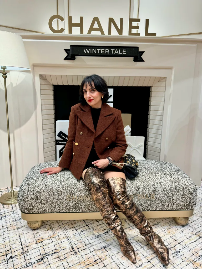 Laura Gallo-Wallis sits in a Chanel store, wearing a brown coat and metallic thigh-high boots.