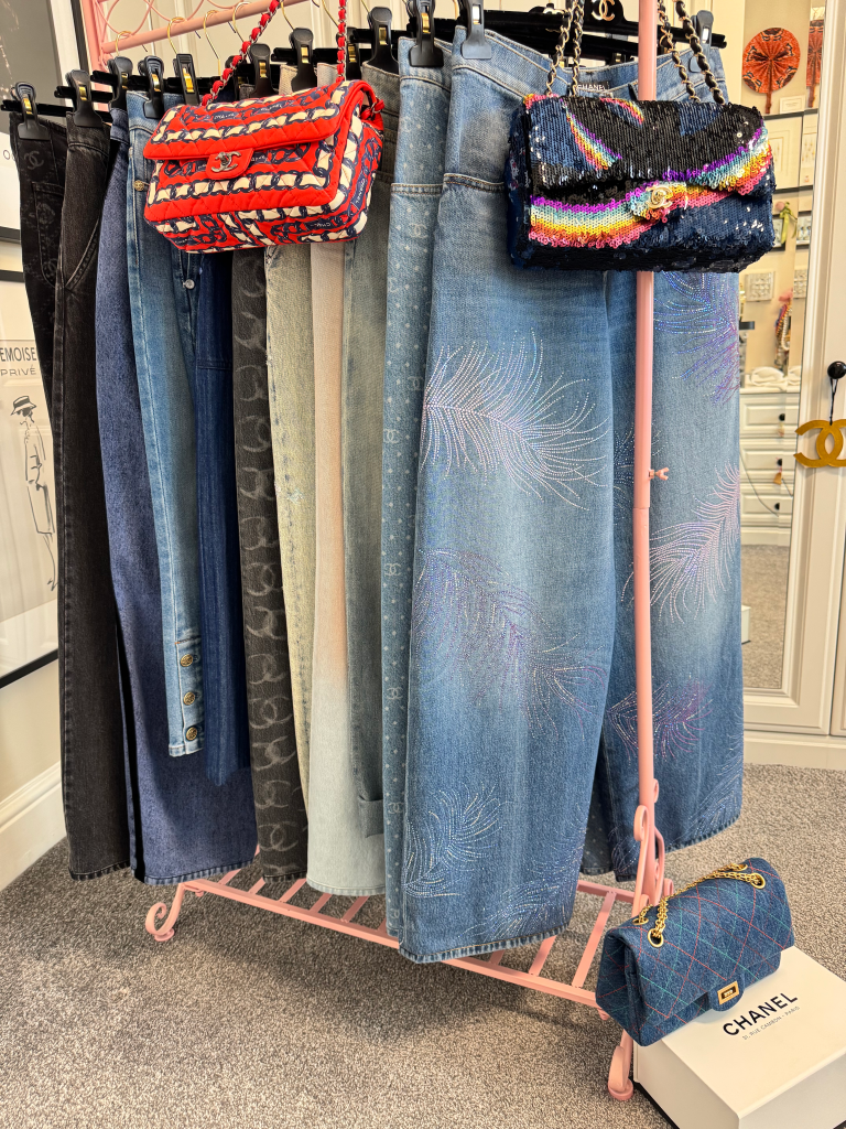 A pink clothing rack holds various Chanel jeans and two Chanel bags, one red, white, and blue, and another black with rainbow sequins, and a denim quilted Chanel bag rests on a box.