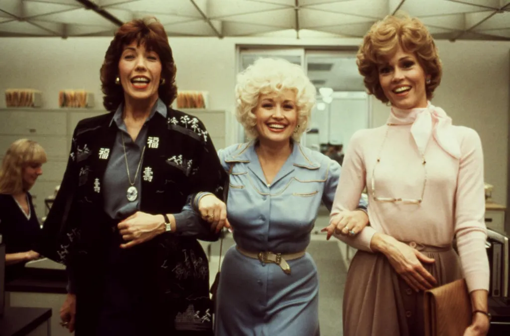 Lily Tomlin, Dolly Parton, and Jane Fonda arm in arm, smiling in an office.