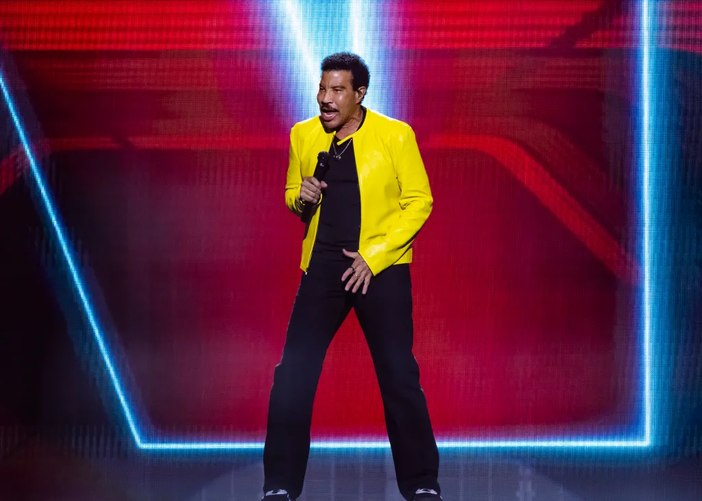 Lionel Richie performing on stage, wearing a yellow jacket and black pants, holding a microphone.