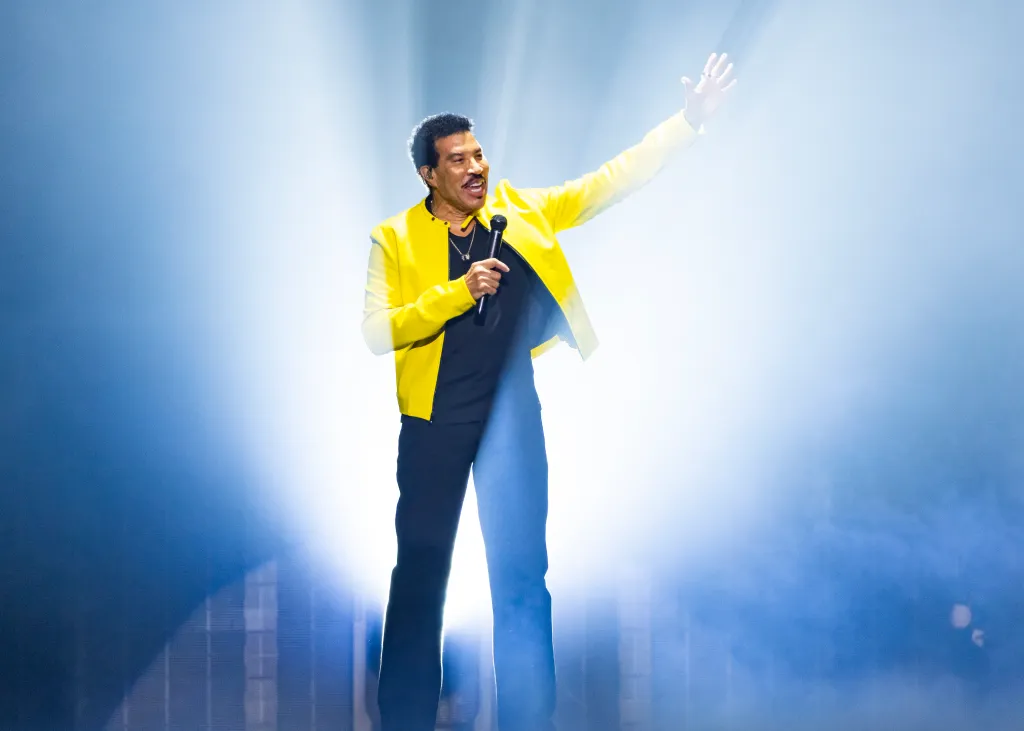 Lionel Richie performing on stage, holding a microphone in his left hand and his right arm raised.