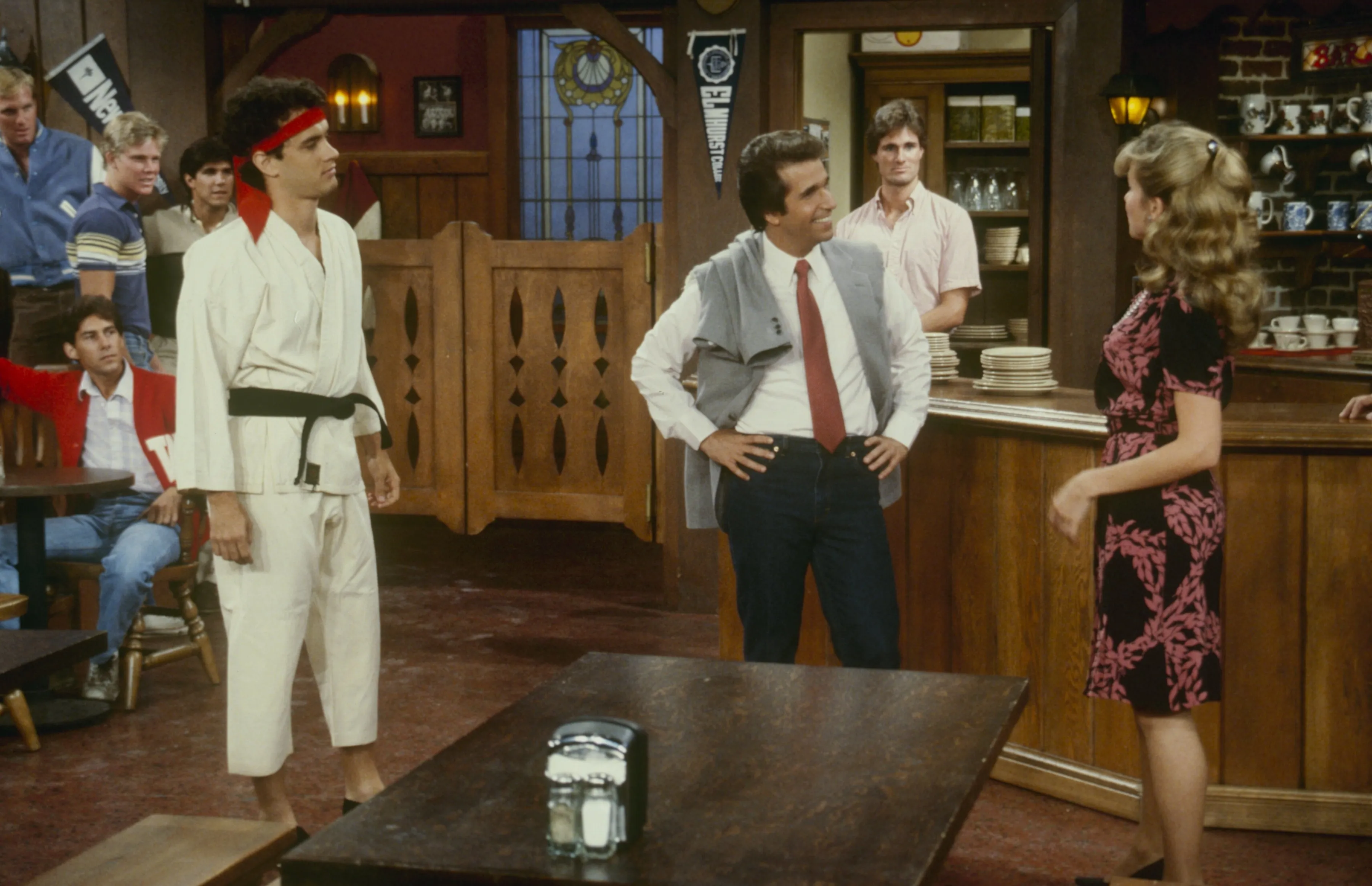 Tom Hanks in a white karate gi, Henry Winkler in a gray vest and red tie, and Linda Purl in a floral dress in a bar scene for 