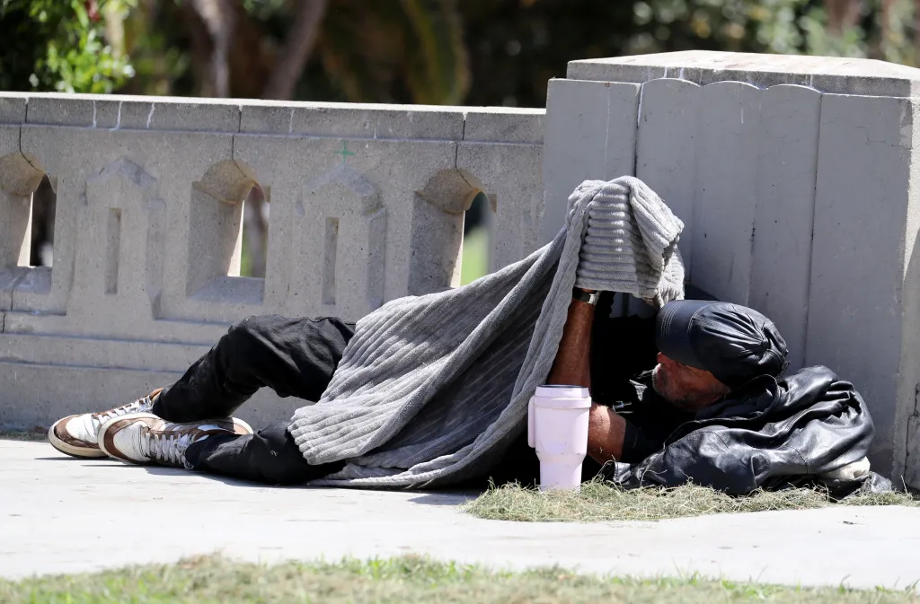 A homeless person lying on the ground next to a concrete wall in MacArthur Park.
