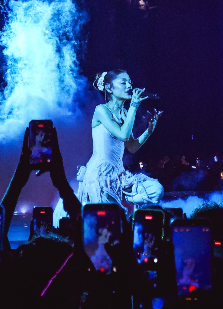Ariana Grande in a white gown performs on stage with blue lights and smoke, while concertgoers film on their phones.