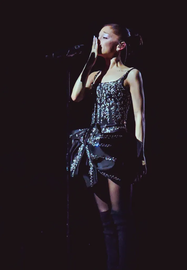 Ariana Grande performing in a dark, sequined mini-dress with a microphone stand beside her.