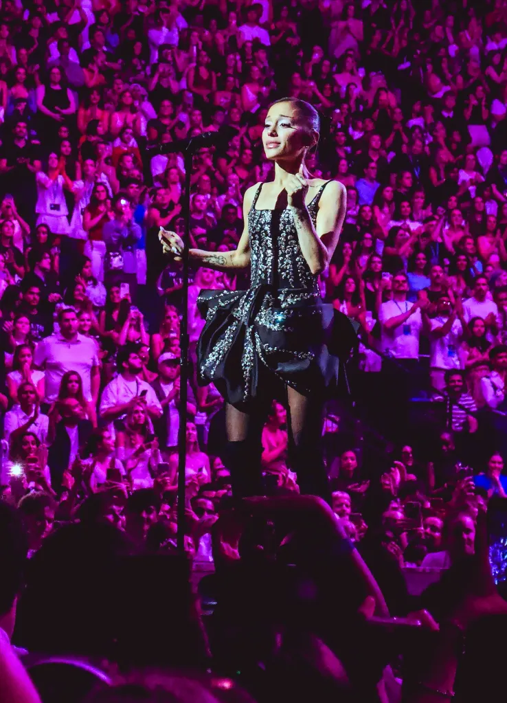 Ariana Grande performing on stage in a sparkly black dress with a large ruffled skirt, surrounded by a cheering crowd.