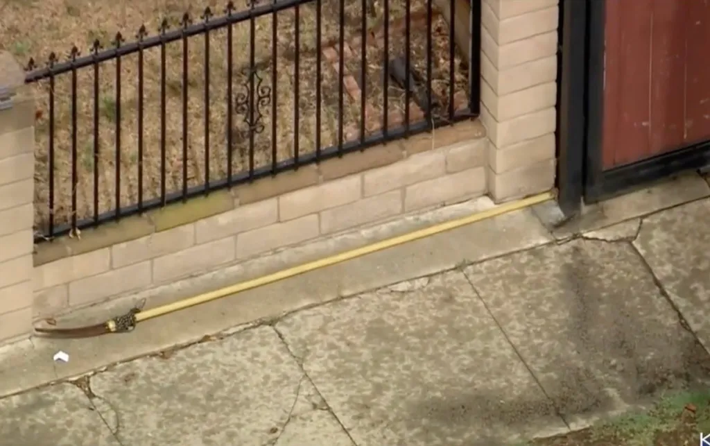 A pole saw with a curved blade and long yellow handle lies on a cracked sidewalk next to a brick wall and black metal fence.
