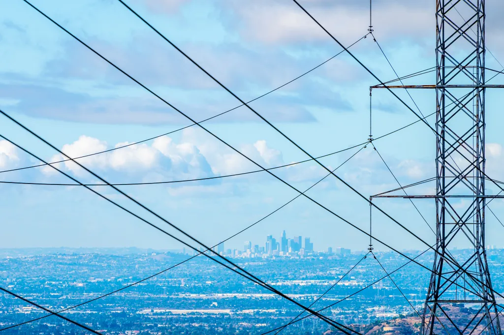 The Los Angeles skyline is seen behind a power tower and power lines on a blue, cloudy day.