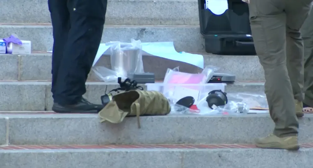 Items spread on stone steps, including what appears to be bomb-making materials and a bag, with parts of two people's legs visible on either side.