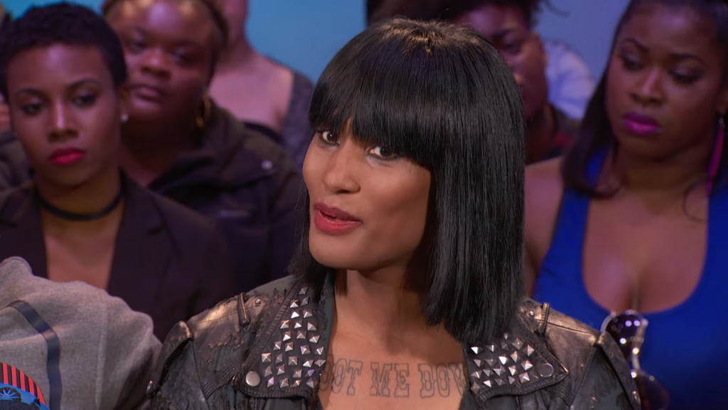 A Black woman with long, dark hair and bangs, wearing a studded leather jacket, with a chest tattoo that says 