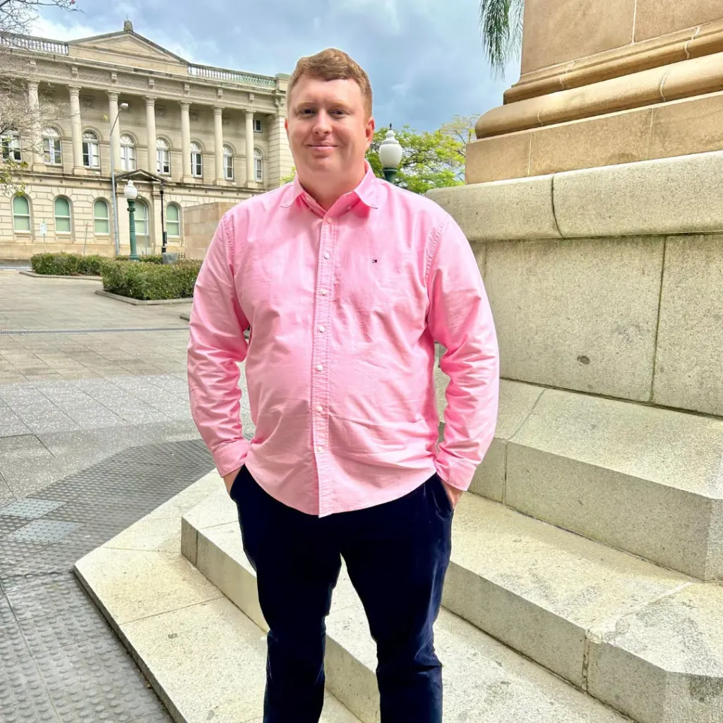 Luke, age 35, wearing a pink shirt and dark pants, standing outside a large building.