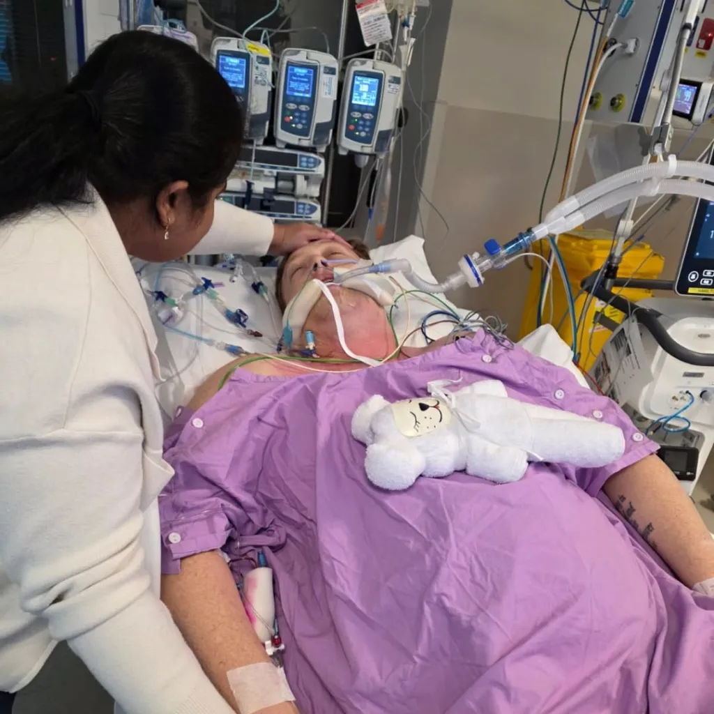 A man in a hospital bed with an intubation tube and tubes in his arms, his forehead being touched by a woman in a white sweater.