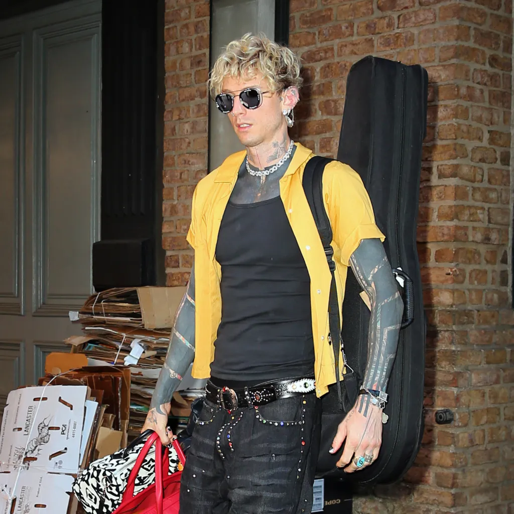 Machine Gun Kelly in a yellow shirt leaving a hotel with a guitar and duffel bag.