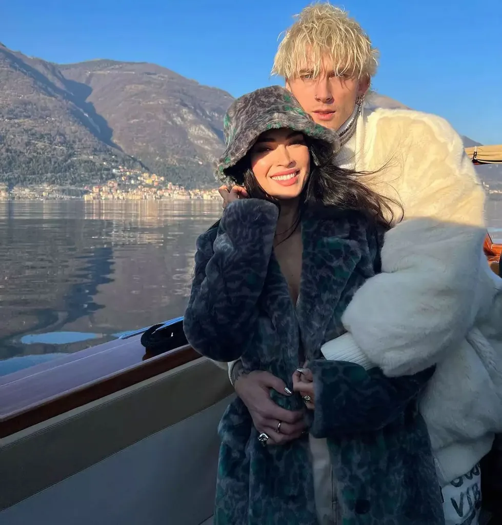 Machine Gun Kelly with his arm around Megan Fox on a boat with mountains and a lake in the background.