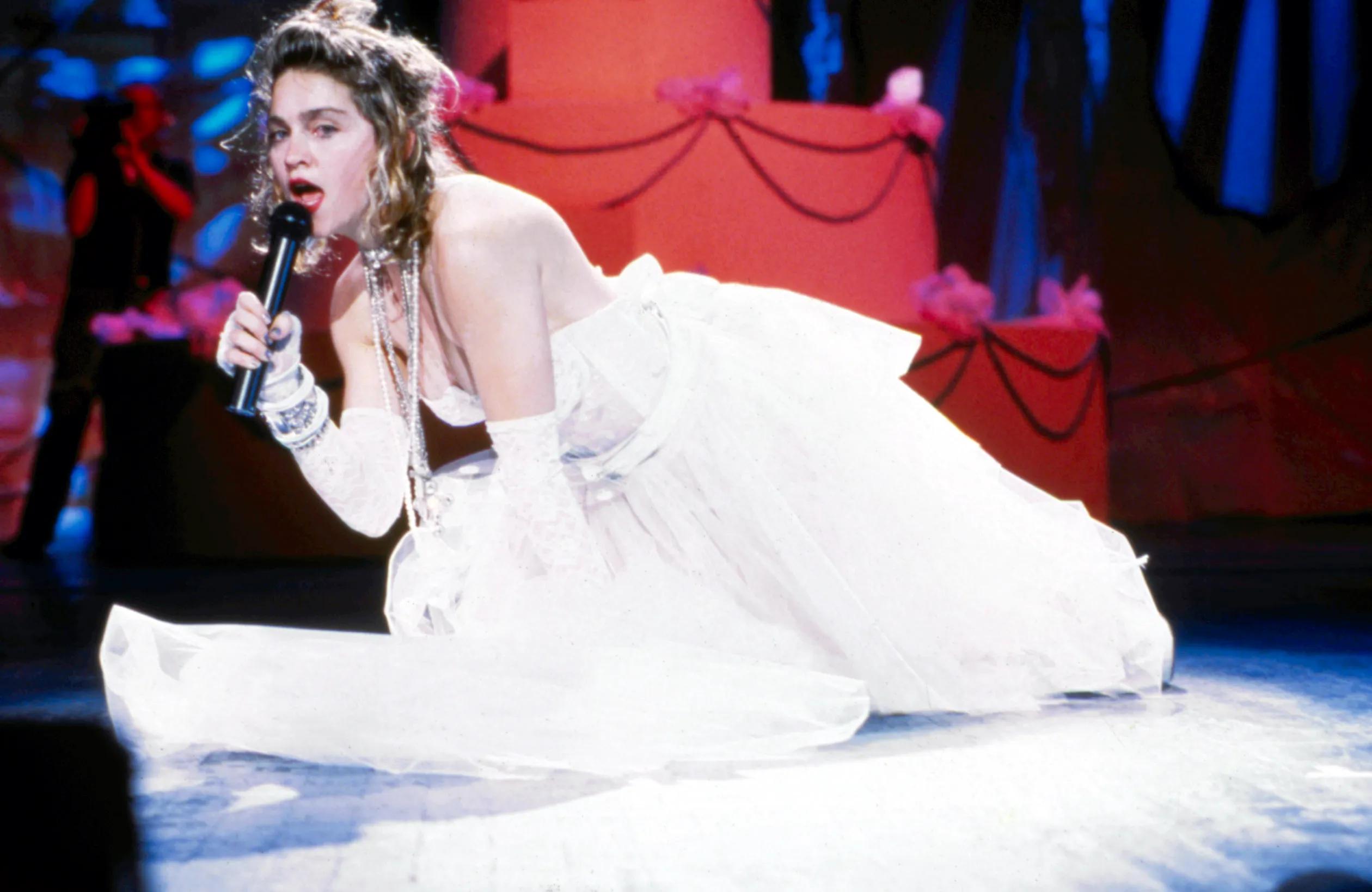 Madonna performing on stage in a white dress and gloves, holding a microphone.