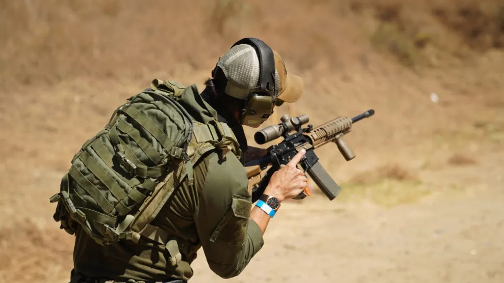 A man in tactical gear with a backpack, hat, and ear protection aims a rifle.