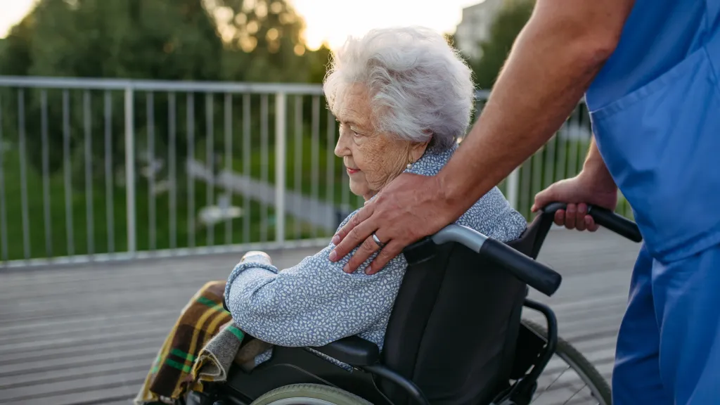 Male caregiver accompanying an elderly patient in a wheelchair on an outdoor walk.