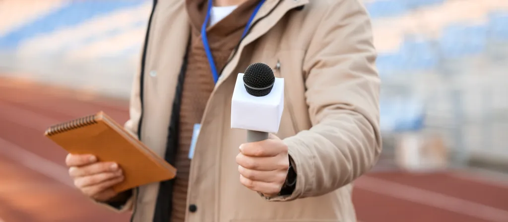 A male reporter holds a microphone and notepad at a stadium.