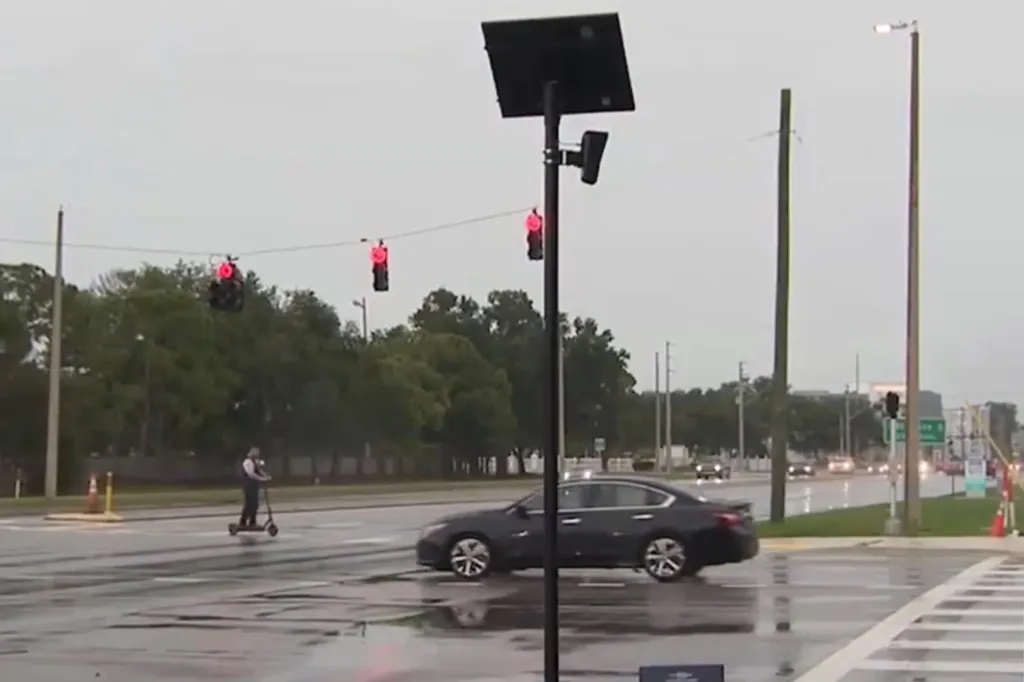 A solar-powered camera mounted on a pole, overlooking a rainy street with a car and a man on a scooter, under red traffic lights.