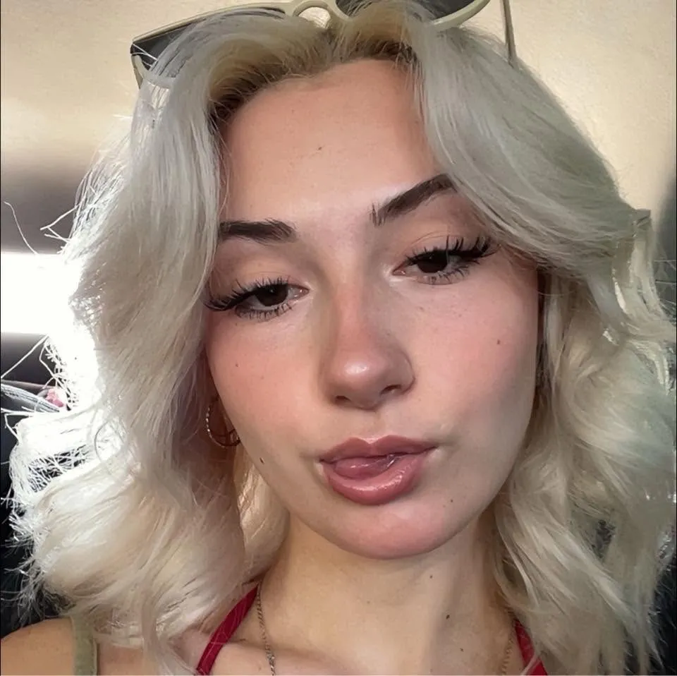 Close-up of CloeRae McBrayer with light blonde hair, sunglasses pushed back on her head, and her tongue sticking out slightly.