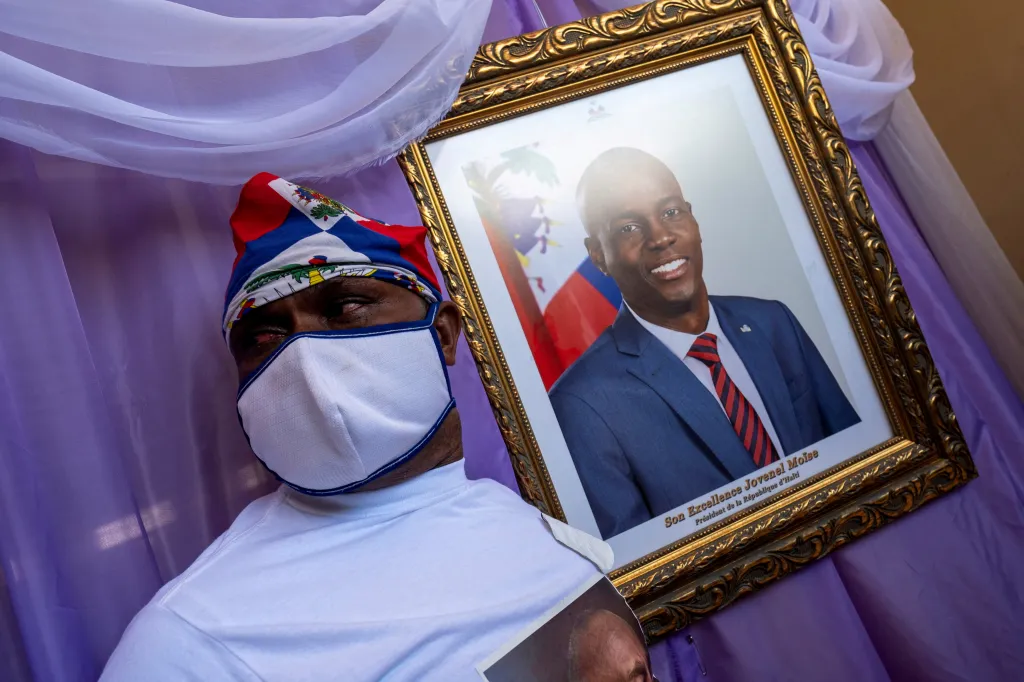 A man with a Haitian flag bandana and face mask stands next to a framed portrait of President Jovenel Moïse.