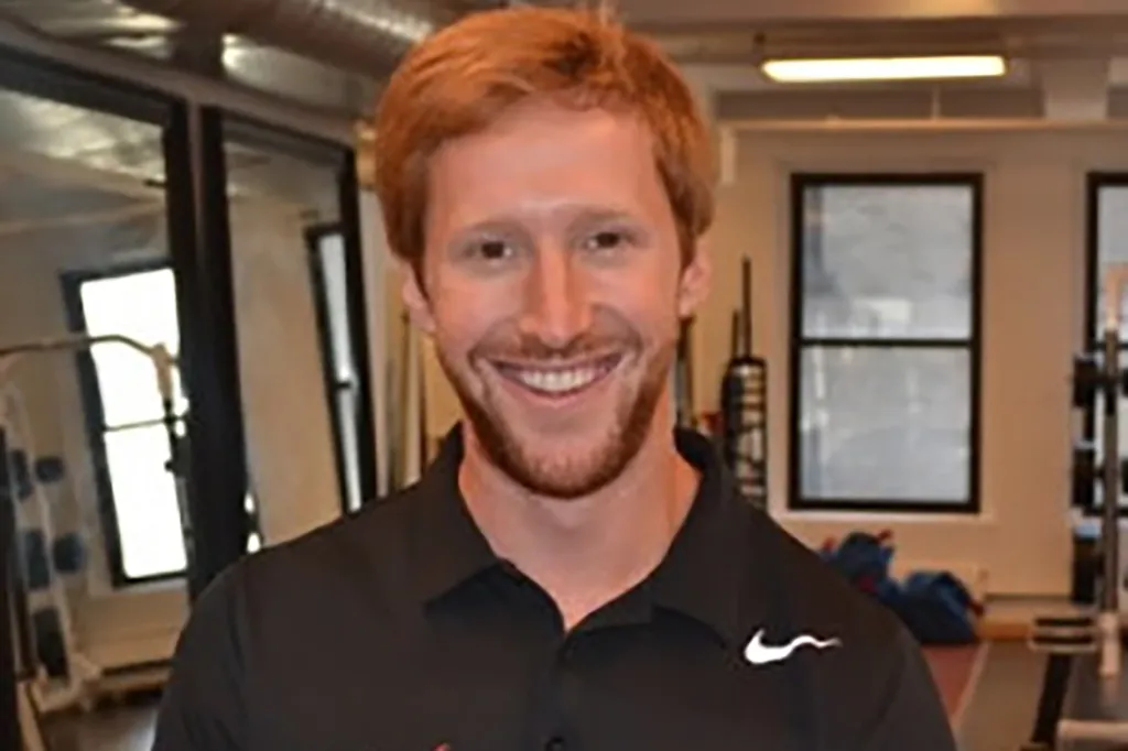 A man with reddish-brown hair and beard smiling, wearing a black collared shirt with a white Nike swoosh.
