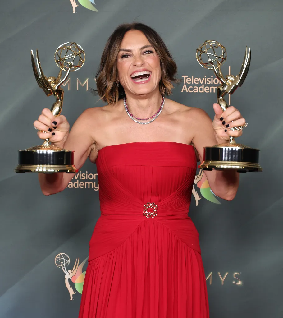 Mariska Hargitay holding two Emmy awards, laughing with her mouth open, wearing a red strapless gown and a ruby and diamond necklace.