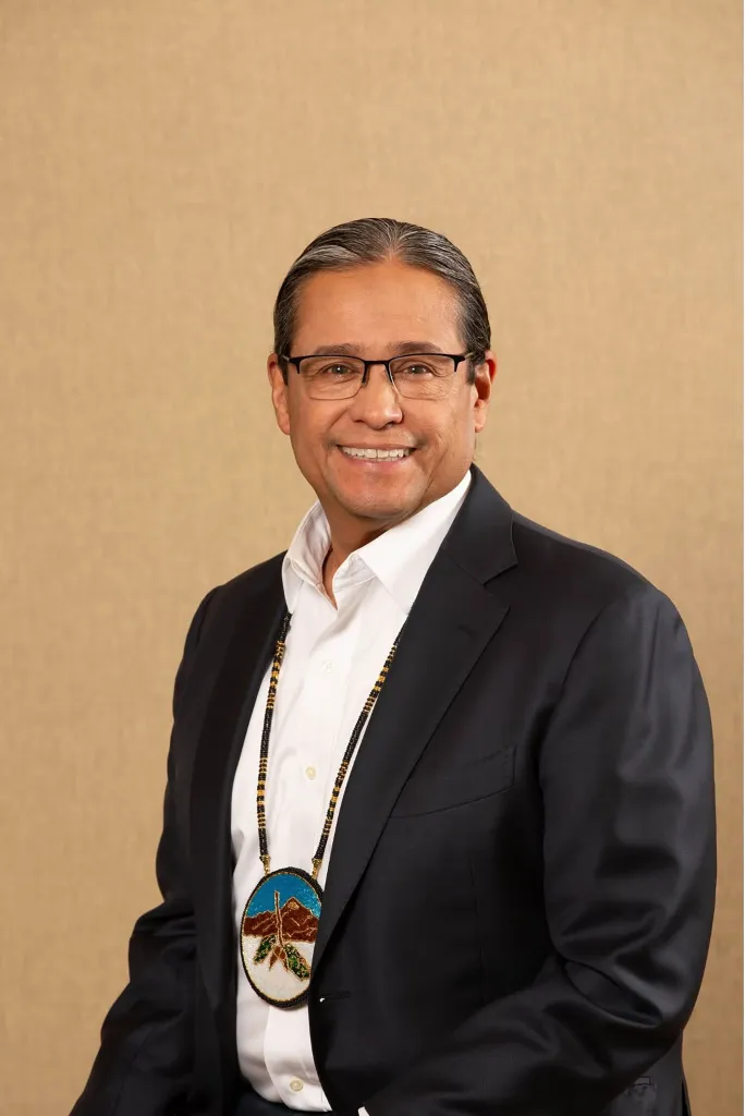 Mark Macarro, president of the National Congress of American Indians and tribal chief of the Pechanga Band of Indians, smiles, wearing a suit and a beaded medallion.