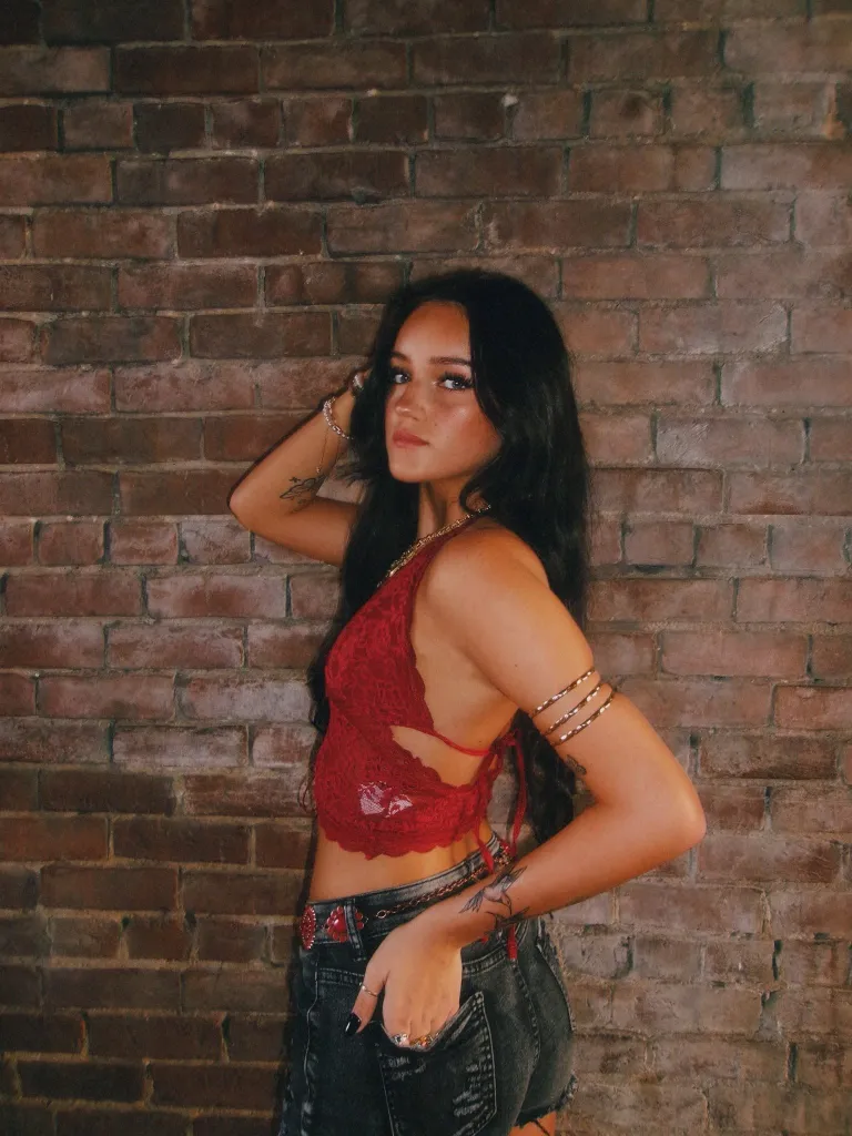 Marly Kinney, a young woman with long dark hair, wearing a red top and denim shorts, posing in front of a brick wall.