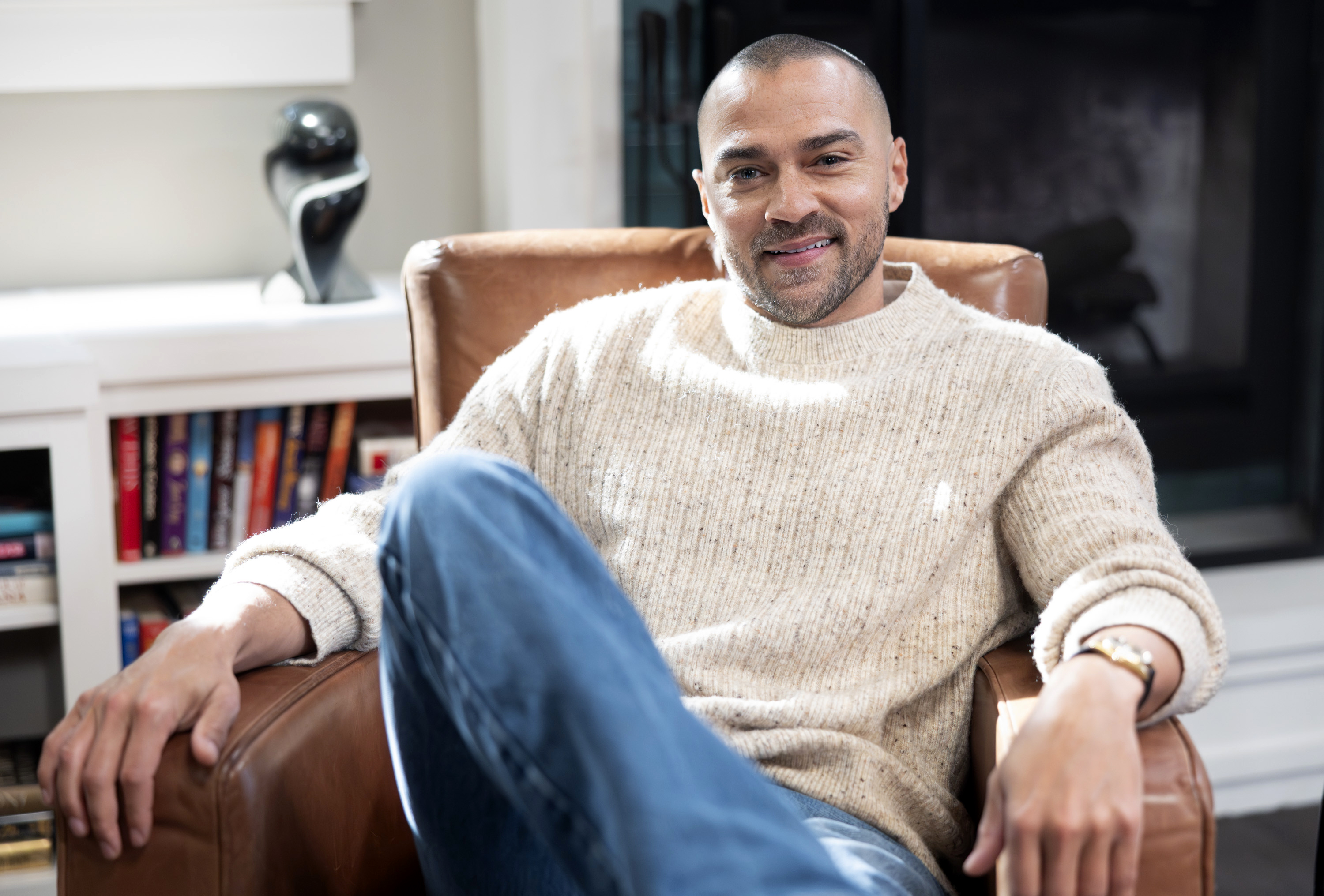 Jesse Williams smiling and seated in a brown armchair.
