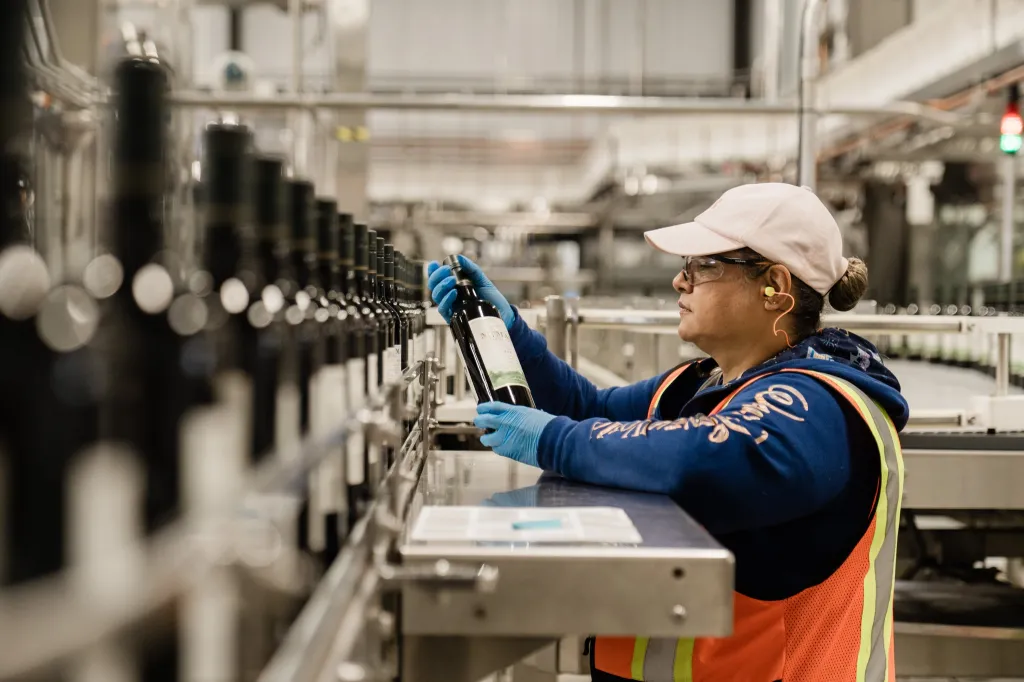 A worker in safety glasses and earplugs inspects a bottle of wine on an assembly line.
