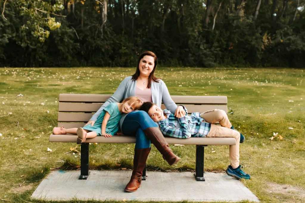 Megan Ireland-Ghekiere sits on a park bench with her son, Owen, and daughter, Chloe, resting on her.