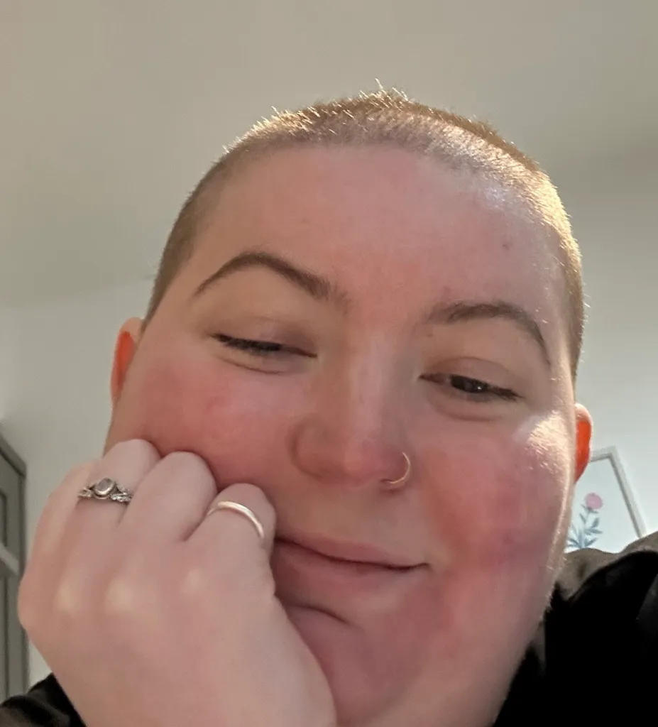 A close-up of Megan Wright, a woman with very short hair and a nose ring, wearing two rings on her left hand and resting her chin on her hand, smiling subtly.