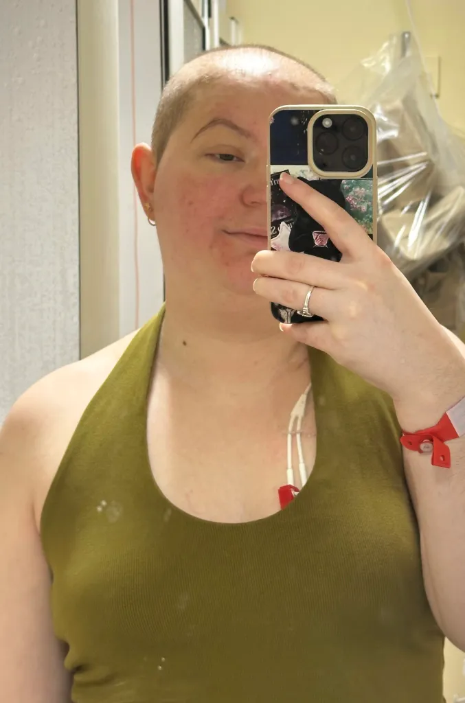 A woman with short hair and a visible port-a-cath in her chest takes a mirror selfie.