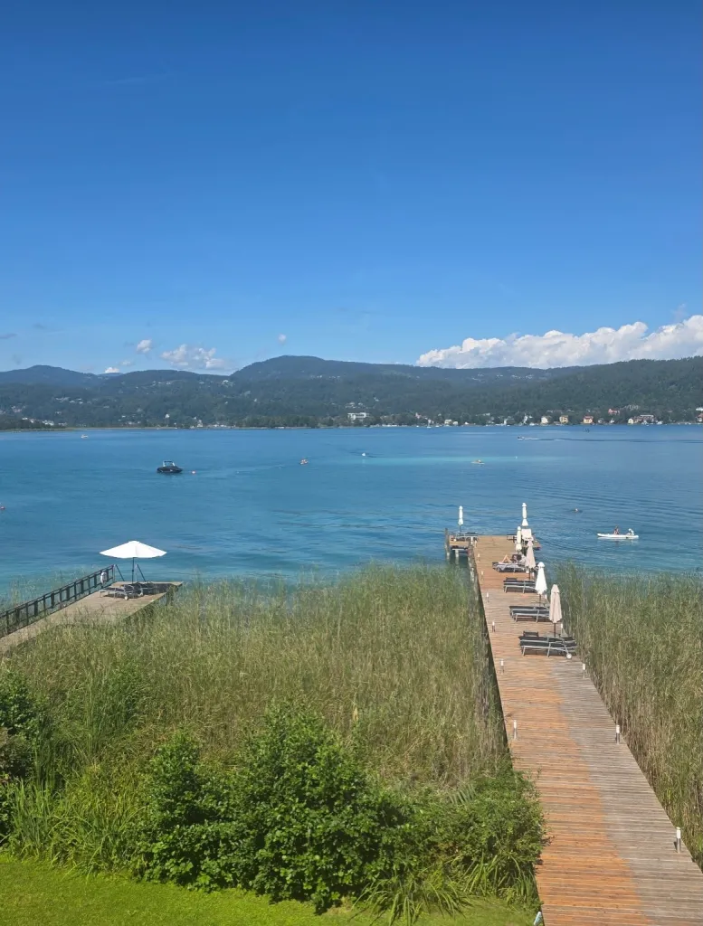 A long wooden pier with lounge chairs and umbrellas extending into a clear blue lake with mountains in the background.