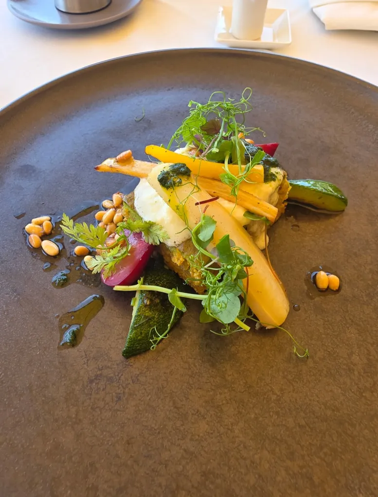 A gourmet dish of vegetables, herbs, and pine nuts on a dark plate.
