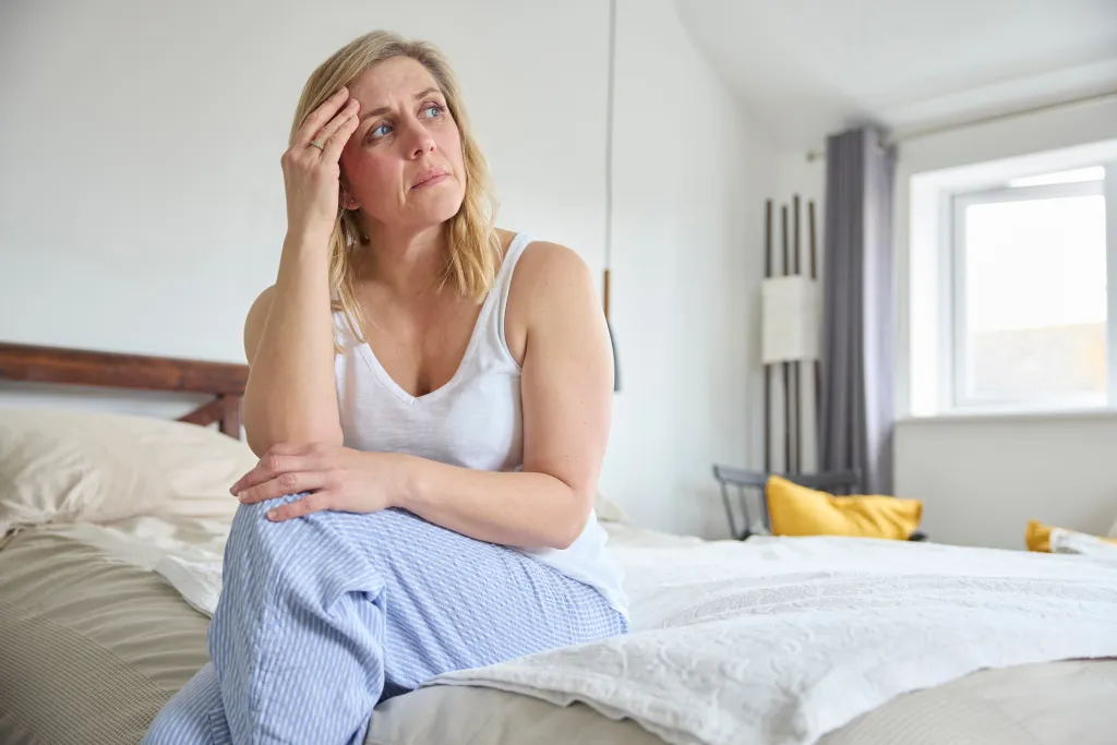 A mature woman in a tank top and pajama pants sits on a bed, appearing distressed as she holds her head and looks up.