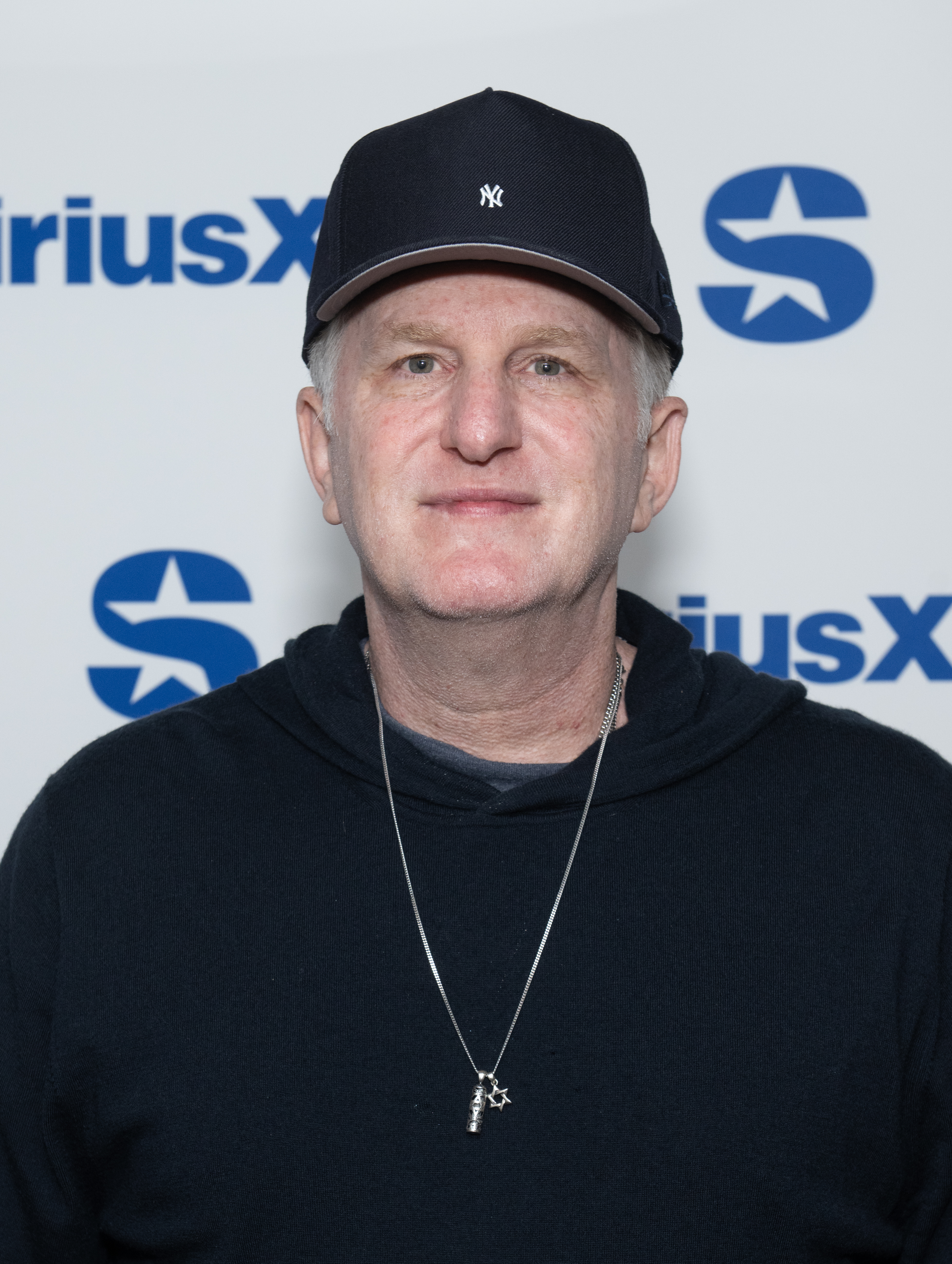Michael Rapaport wearing a New York Yankees baseball cap and a black hoodie with a Star of David pendant, standing in front of a SiriusXM Studios backdrop.
